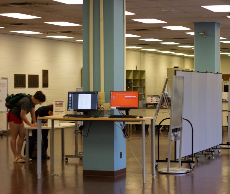 A spacious room with a drop ceiling and fluorescent lights. Two students are bent over a small table. Another higher table has two computer monitors. Photo by Jorge Murga.