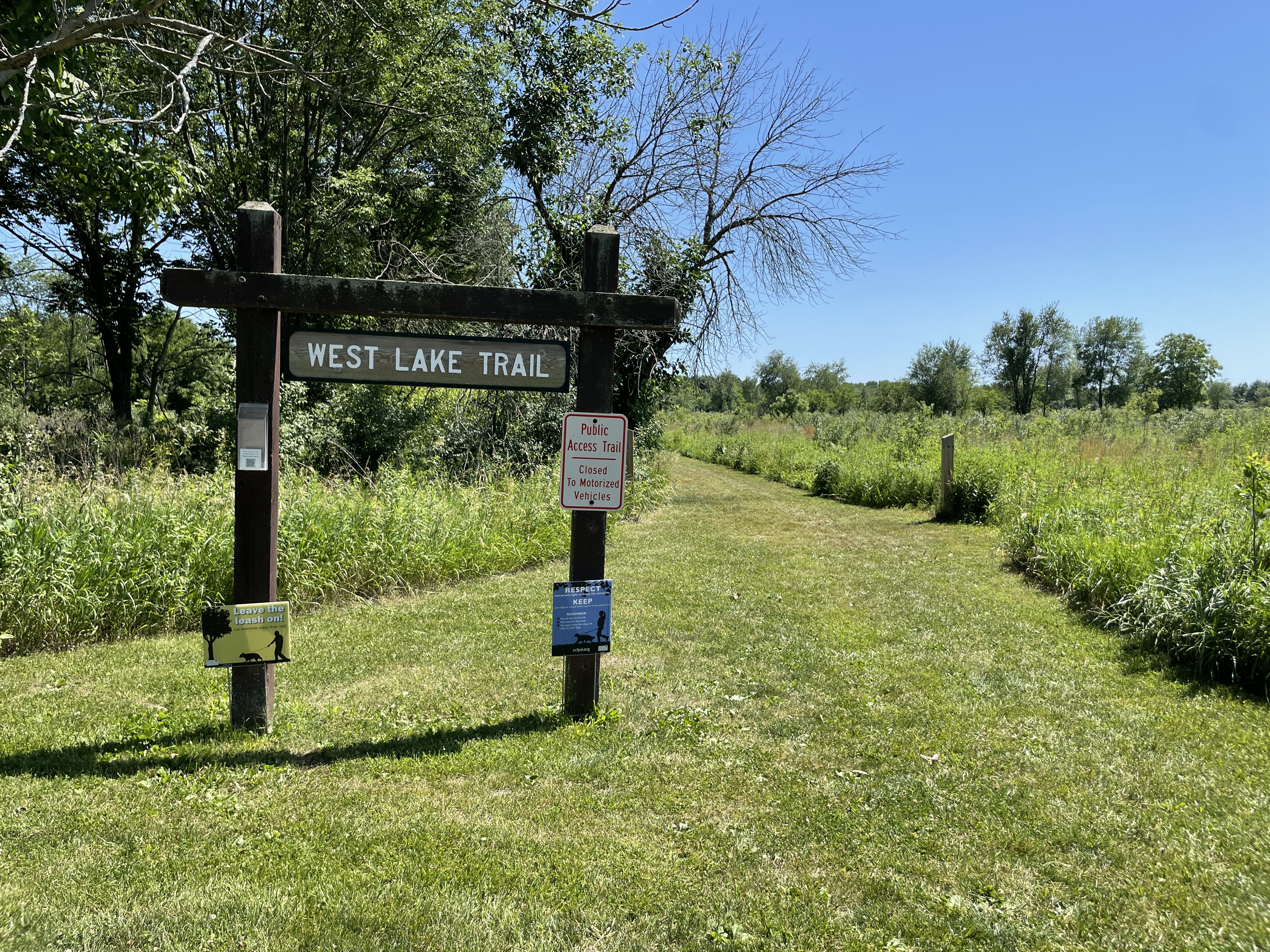 A photo of the West Lake Trail trailhead sign with the prairie and some trees in the background. Main colors are greens and blue sky in the back.