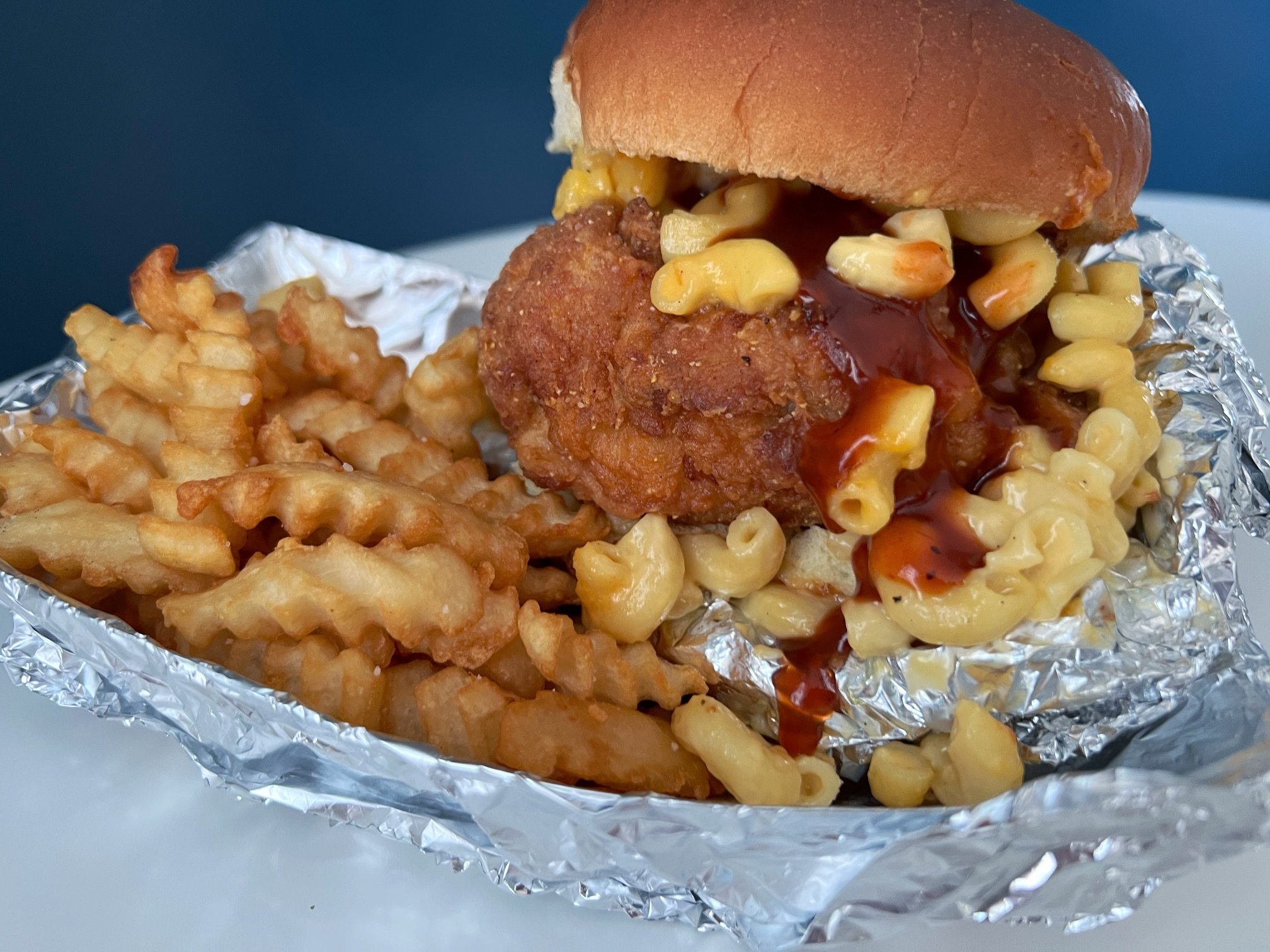 In a tin-foil lined basket, there is a fried chicken sandwich with macaroni and drippy hot sauce on a bed of crinkle fries from The Stuft Bird in Champaign, Illinois. Photo by Alyssa Buckley.