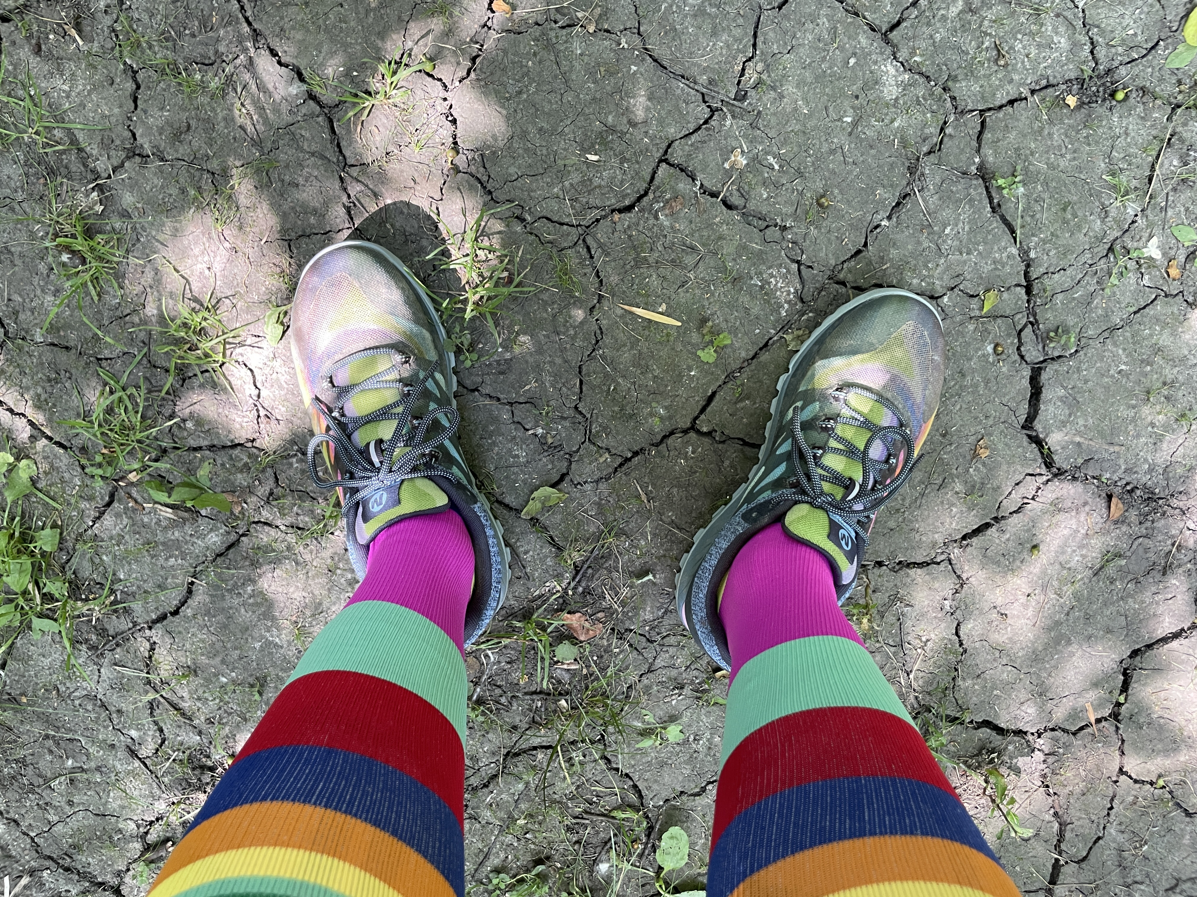 Photo of legs in colorful hiking shoes and socks standing on dry, cracked, brown dirt.