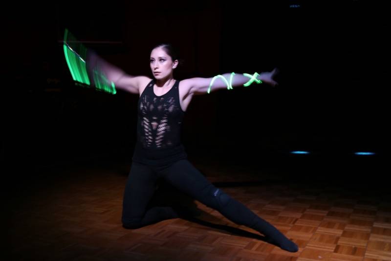 A performer on a stage. She is dressed in all black, and kneeling on one leg with the other outstretched. Her arms are outstretched with green glowing ropes wrapped around them. Photo by Candie Kates Photography.