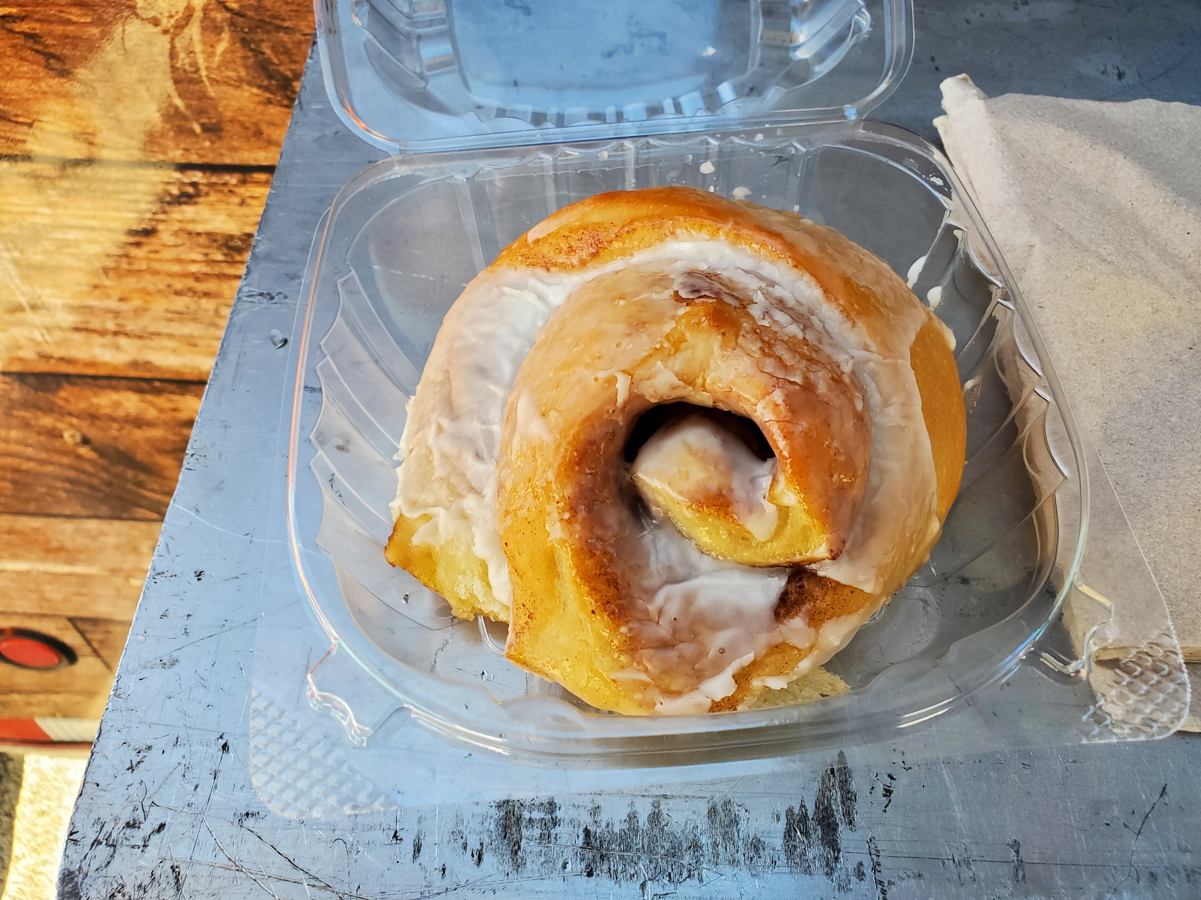 In a clear plastic container, there is a large cinnamon roll with a crackly glaze. Top image by Carl Busch.