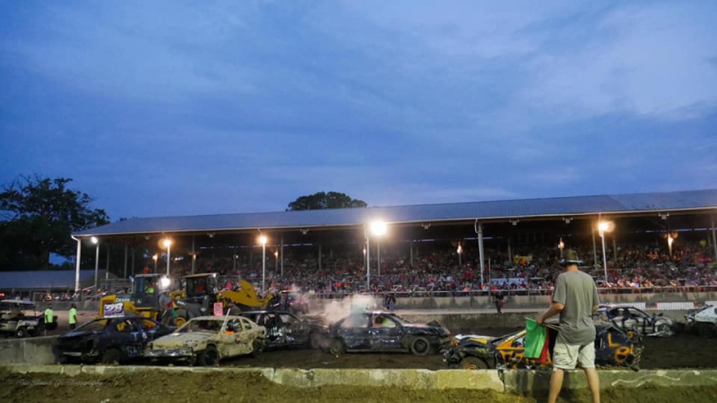 A dirt arena filled with several beat up cars, driving into each other. There is a grandstand filled with spectators in the background. Photo from Champaign County Fair Facebook page.