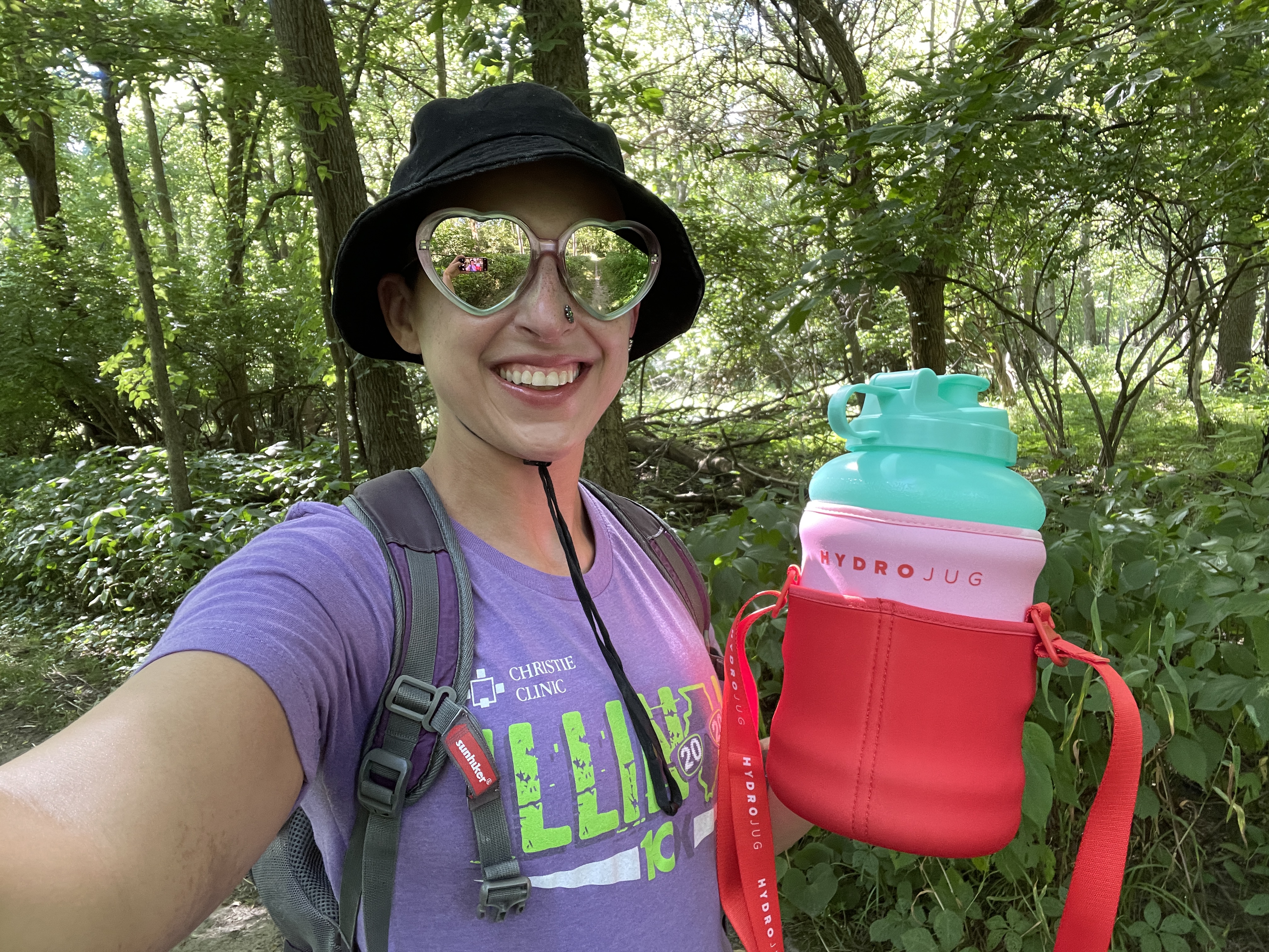 Photo of author Mara Thacker standing with her gallon jug of water and woods in the background. Thacker is wearing sunglasses. Main colors are greens and browns.