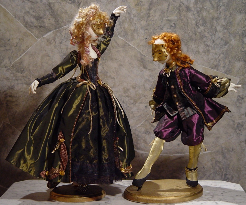 An image of two of Barbara Schoenoffâ€™s dolls, entitled, Venetian Dancers. The dolls are posed as if in mid-dance, each mounted on a small, round, platform. The dolls wear masks that cover the top half of their faces, showing that they are at a masked ball. The period of the dancers is the 1770s.