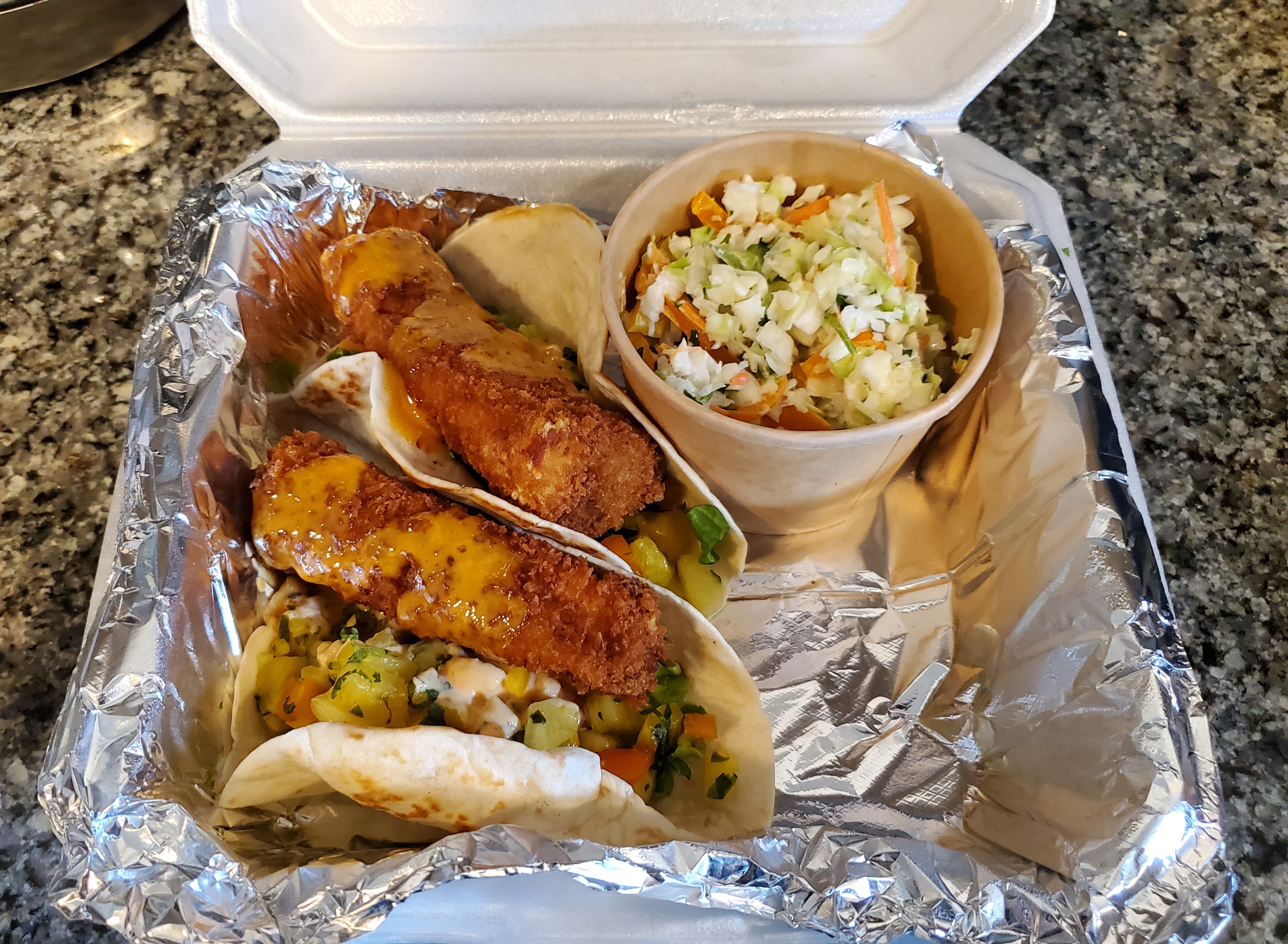 In a tin-foiled to go container, there are three fish tacos with crispy fried fish in flour tortillas with a cup of coleslaw. Top image by Carl Busch.