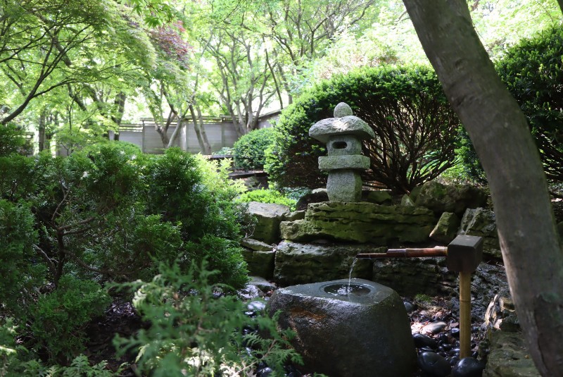 A garden at Japan House with a stone statue and water feature in the center. They are surrounded by green leafy bushes and trees. Photo from Japan House Facebook page.