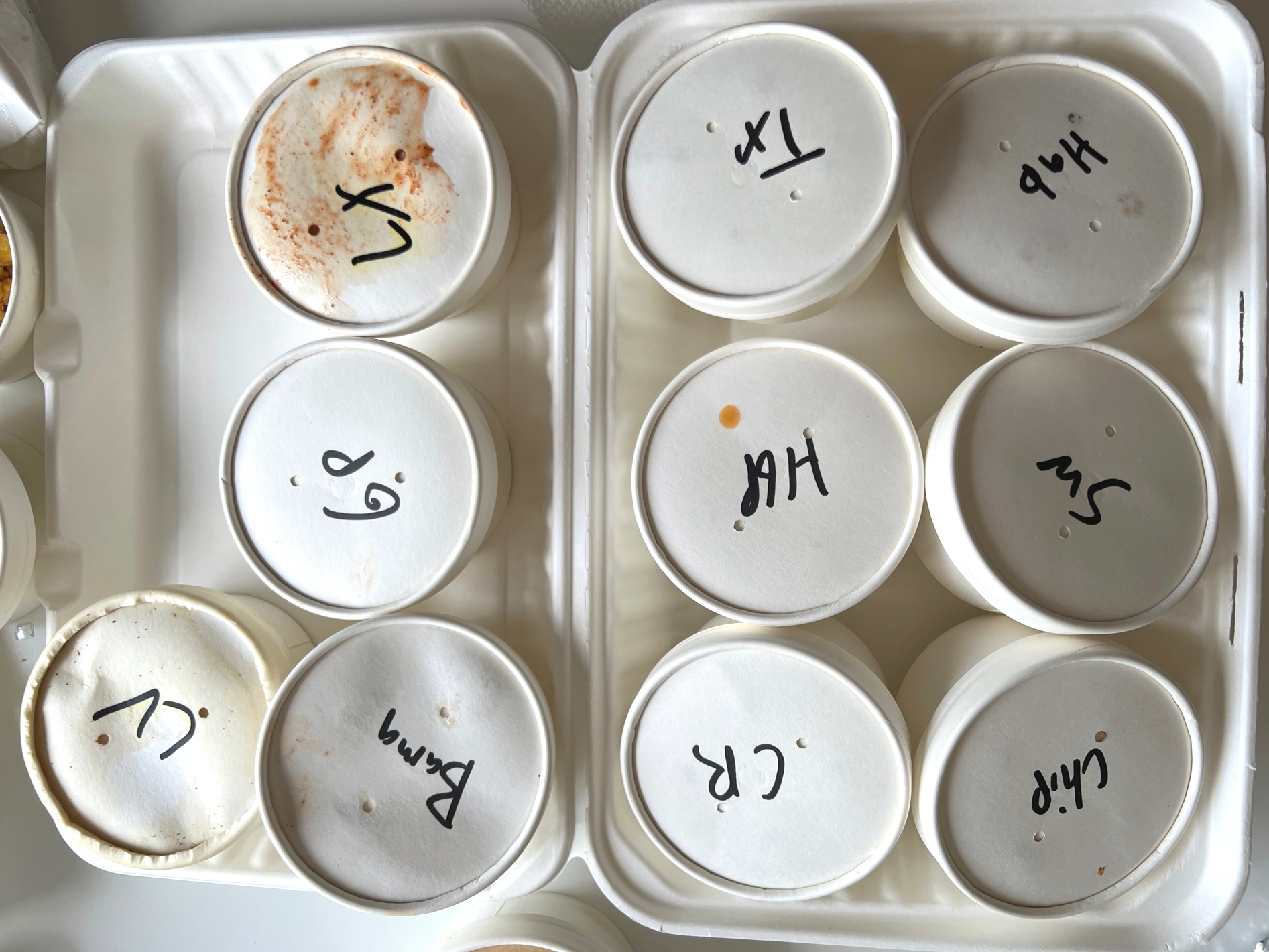 In a white styrofoam container, there are ten white lidded cups with barbecue sauce from Black Dog Smoke and Ale House in Champaign. Each cup has a shortened version of the sauce's name written in Sharpie. Photo by Alyssa Buckley.