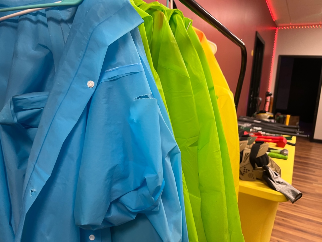 Colorful ponchos of blue, green, and yellow hang on a small rack at Rage Room Champaign on the left side of the photo. Behind the hanging ponchos, there is a yellow table with helmets and weapons. Photo by Alyssa Buckley.