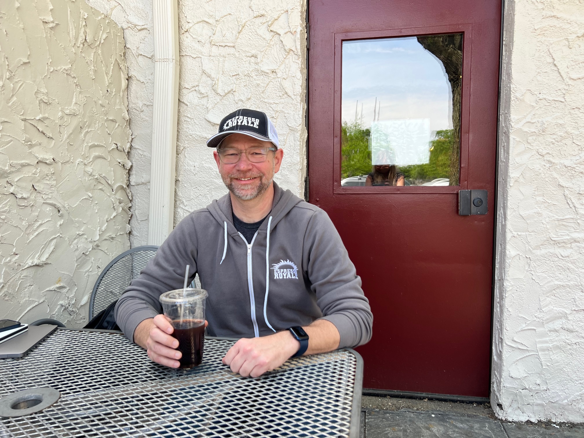 Owner of Cafe Kopi and Espresso Royale Douglas McCarver sits at an outdoor table at the Espresso Royale Krannert View holding a cup of cold brew in a pastic cup with a plastic straw. He is wearing a black hat and gray sweatshirt with Espresso Royale Coffee on it. He is smiling for the camera. Photo by Alyssa Buckley.