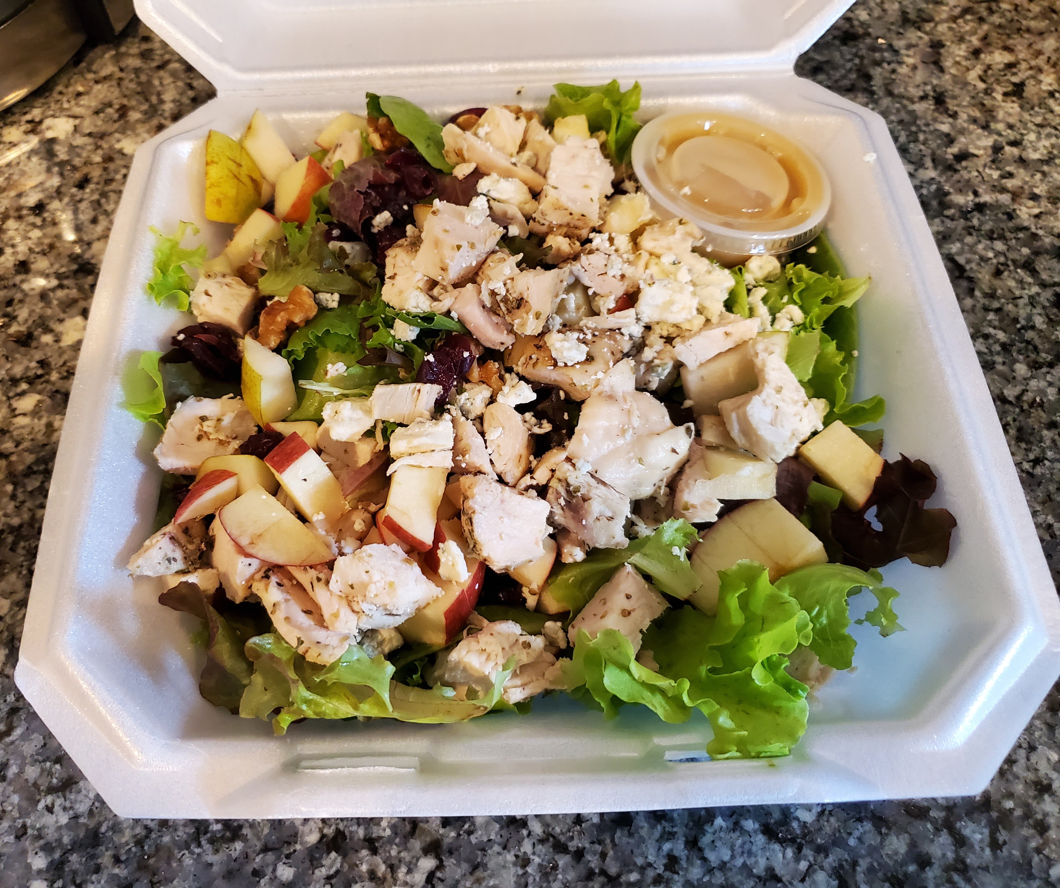 In a styrofoam container, there is a large salad with chicken, cheese, and fruit. Top image by Carl Busch.
