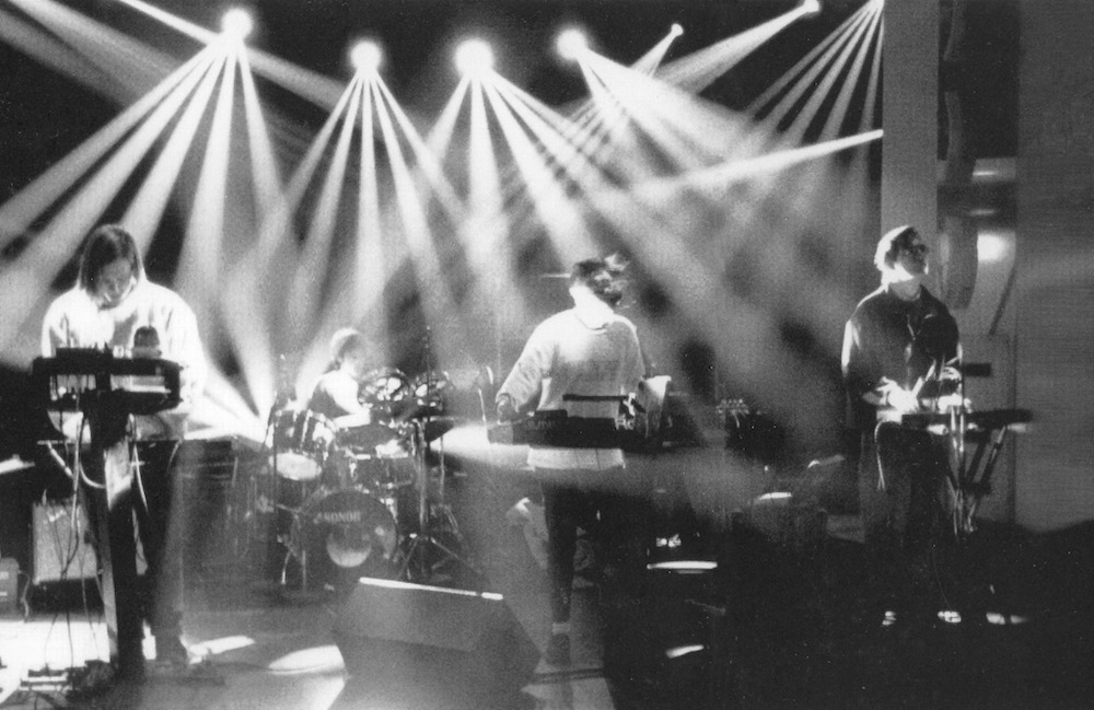 Grainy black and white image of Salaryman performing on stage. Lights overheads shine down on the band.