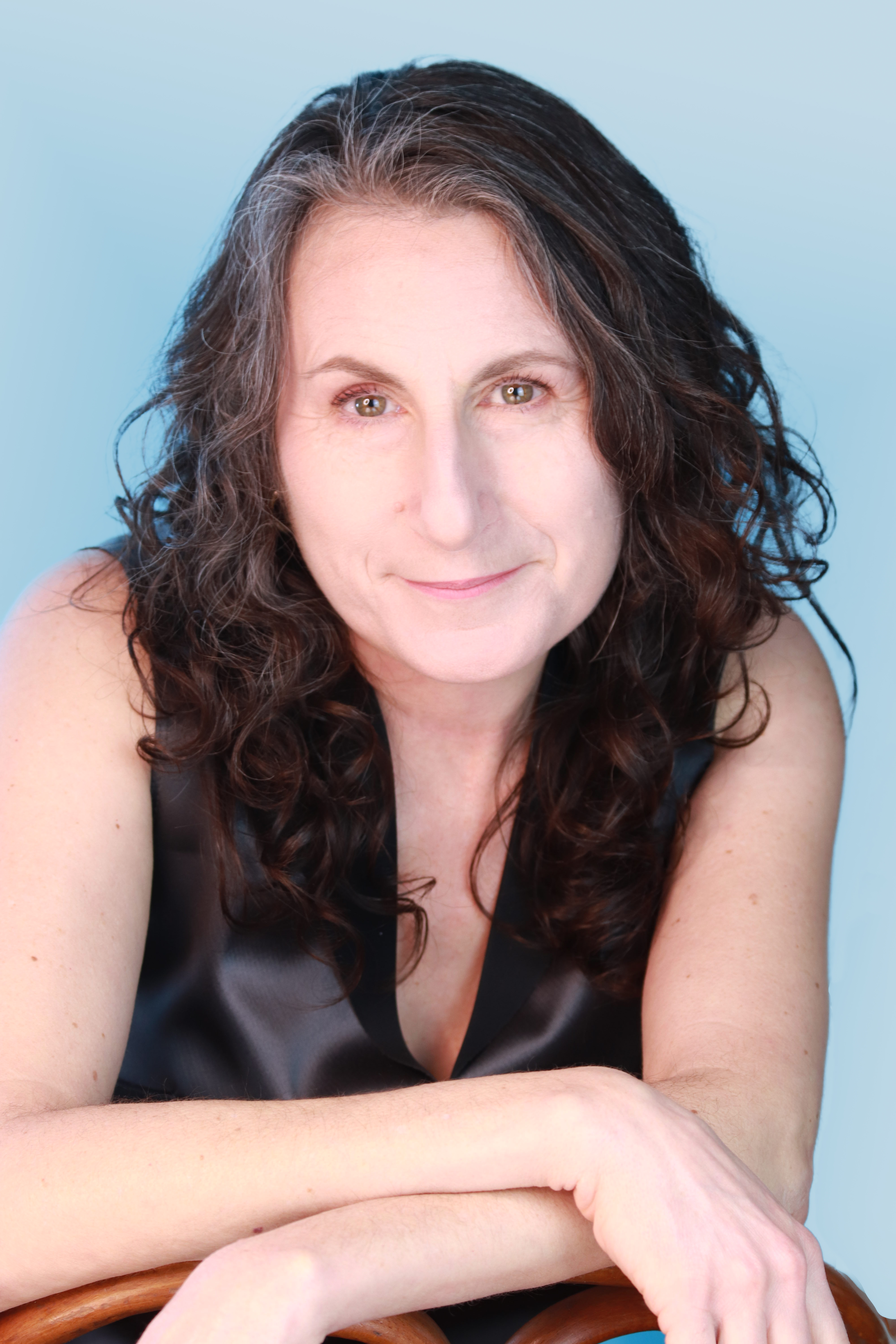 Portrait of Brett Ashley Kaplan. She is a white woman with dark hair with some gray on the part. She is wearing a black sleeveless shirt and looking into the camera. Her arms are folded across the back of a chair. The background is light blue. Photo courtesy of Brett Ashley Kaplan. 