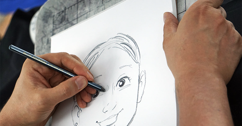 Close-up photo of Dan wild drawing a caricature. His hand is holding a pencil to a white piece of paper as he adds detail to a drawing of a face.Photo from Dan Wild's Facebook page.