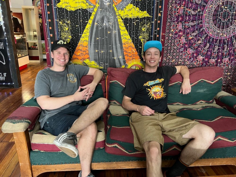 Two white men in baseball caps, t-shirts, and shorts are sitting on a couch, with a tapestry and lights hanging behind them. Photo by Julie McClure.
