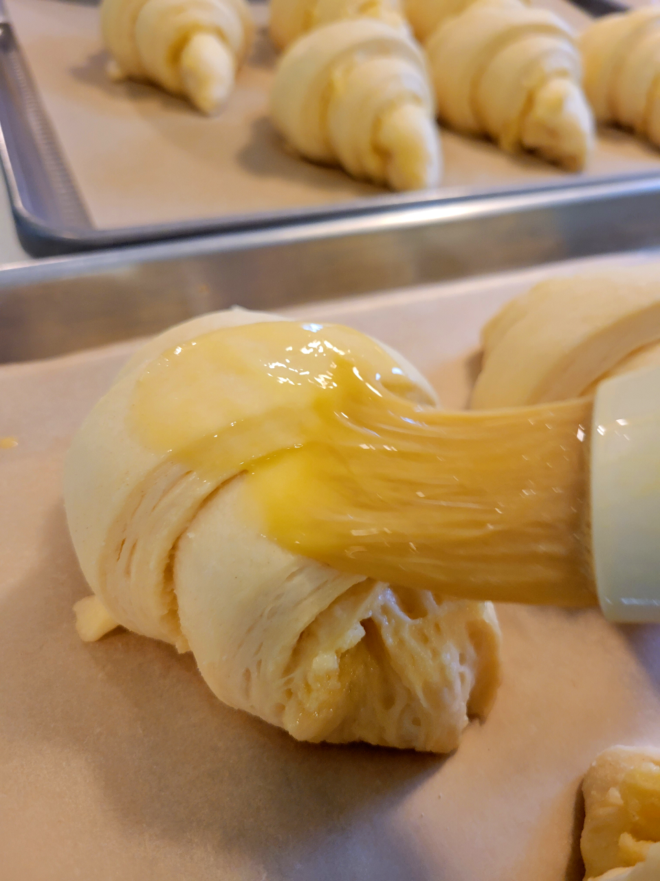 An unbaked croissant on a parchment-lined baking sheet is being brushed with butter. Several other unbaked croissants on a different baking sheet can be seen in the background. Photo courtesy of Kat Fuenty.