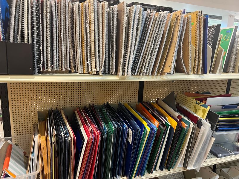 A set of metal shelves with a row of spiral bound notebooks on the top shelf and a row of two pocket folders on the bottom shelf. Photo by Julie McClure.