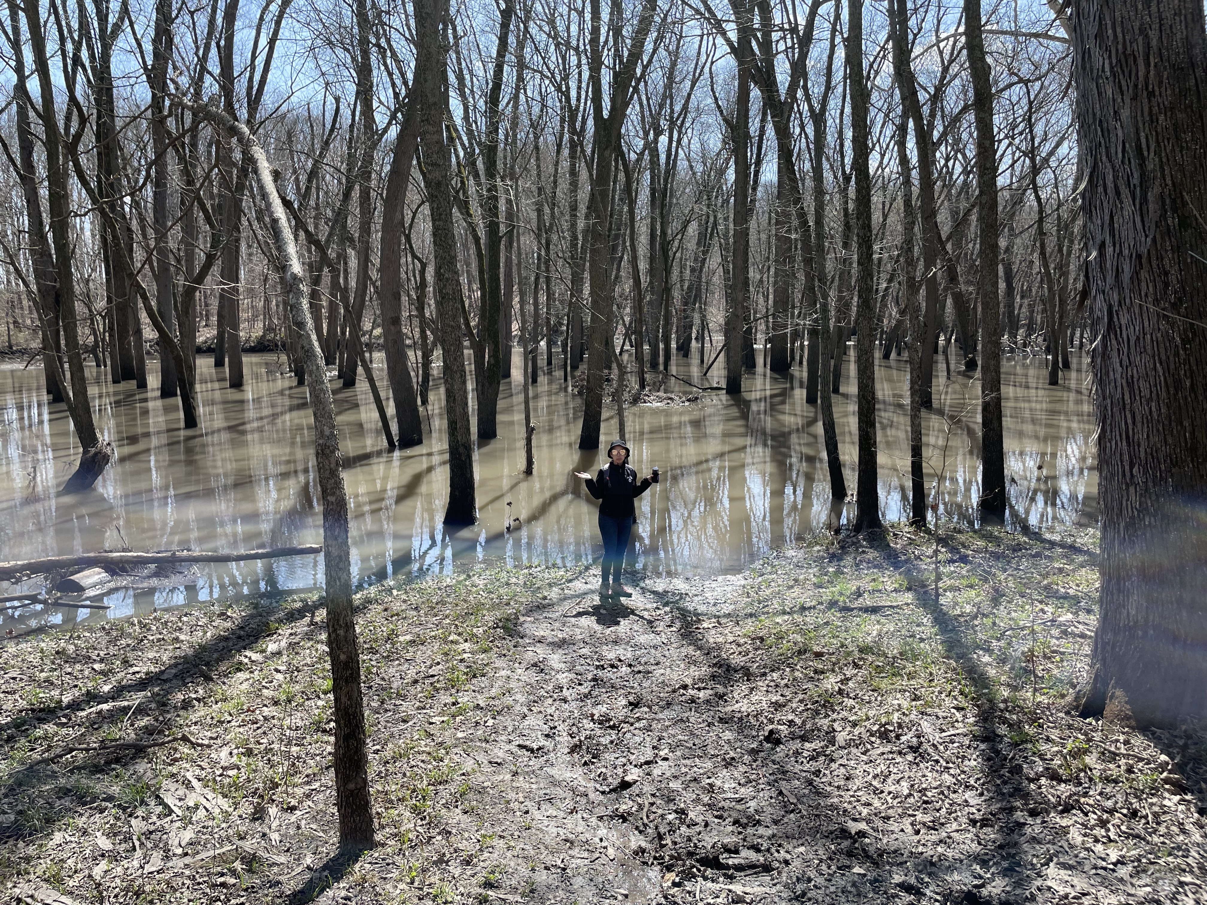 Photo of Mara standing in front of a murky body of water where the trail was intended to continue. Main colors are browns. This photo is from April 2022 whereas the other photos are from August 2022.