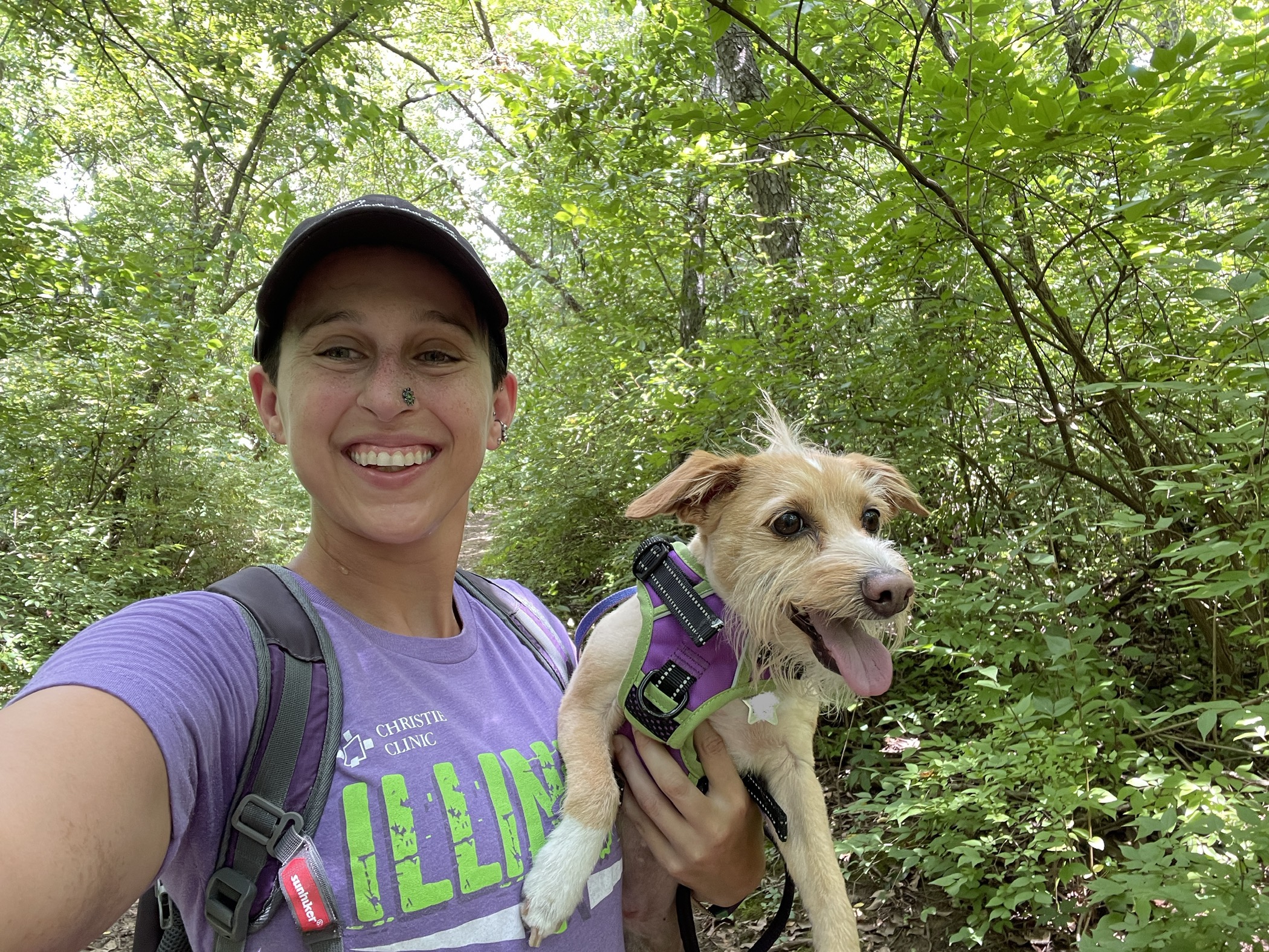Photo of the article's author standing in front of the trail holding her dog. Mara is wearing a purple t-shirt and the dog is small, beige, and scruffy with his tongue out. There are trees in the background. Main background colors are brown and green.