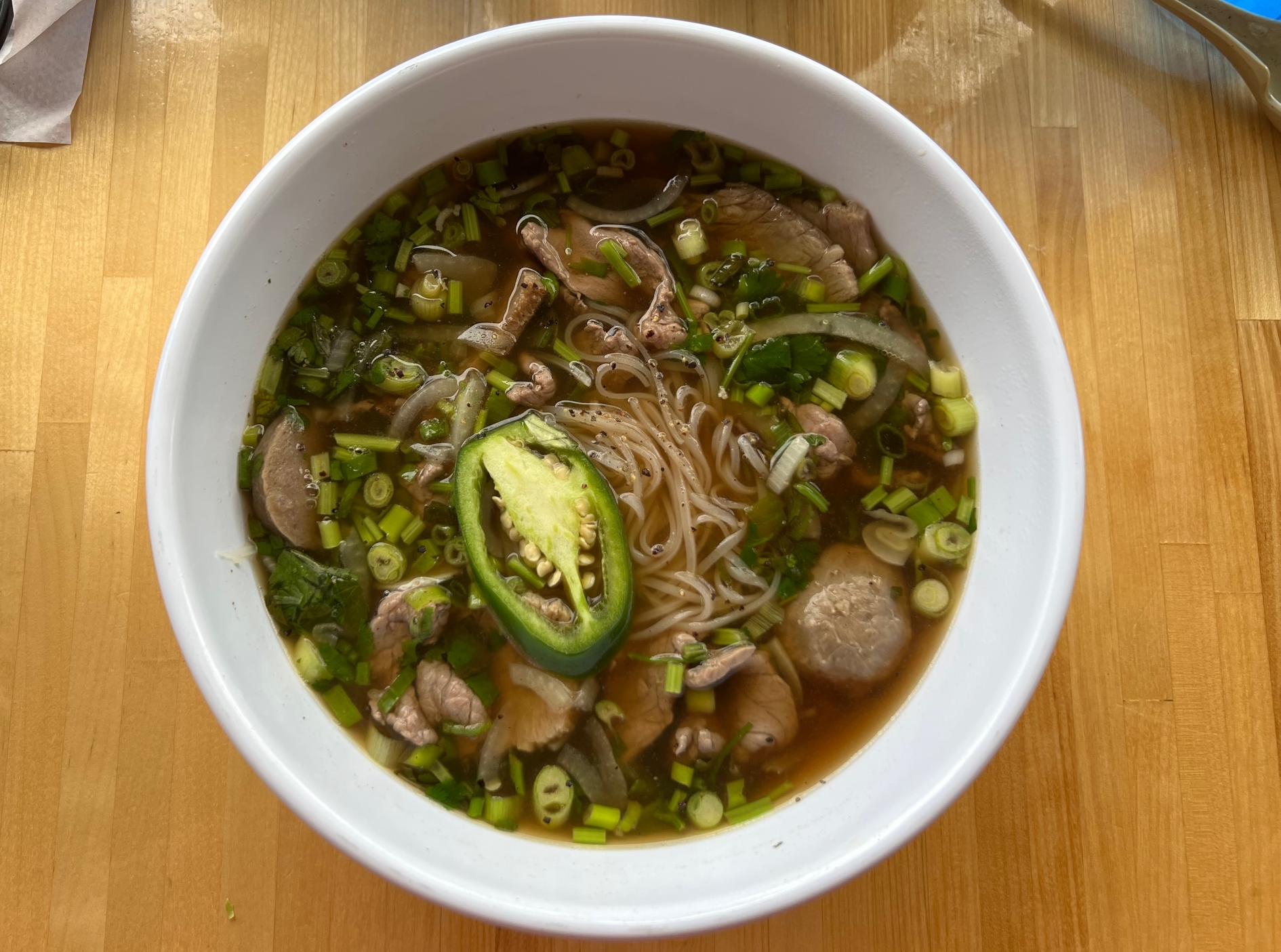 On a wooden table, there is a white bowl of pho with jalapeno, beef, meatballs, green onion, and rice noodles. Photo by Alyssa Buckley.