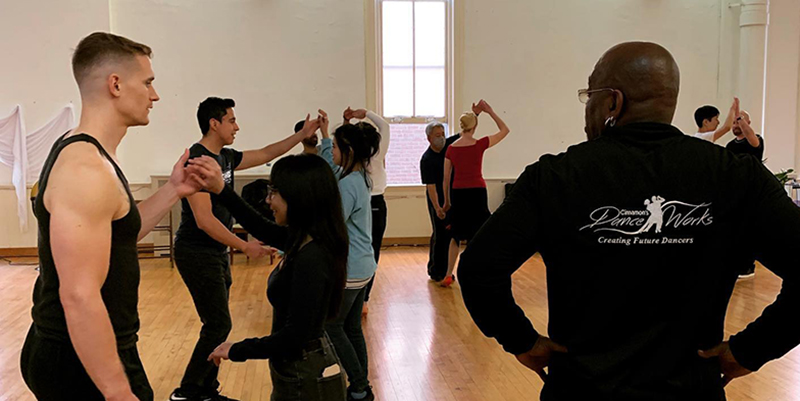 Several dance duos practicing Latin dance moves in a white walled dance studio with wood floor. Up front are two male instructors looking on.