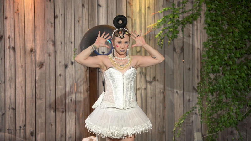 A performer in a white corset and white tulle skirt with a black donut shaped headband posing. Photo by Jeff Putney.
