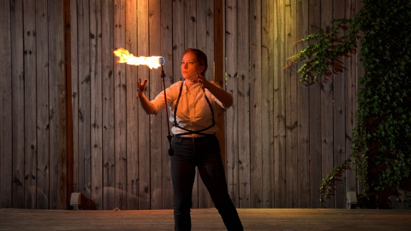A woman in a white shirt and black pants with thin black suspenders is playing with a firestick that appears to be suspended in mid-air. Photo by Jeff Putney.