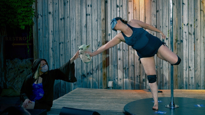 A  pole dancer in a mask, black tank top and shorts, and black kneepads handing off a toy of Baby Yoda to a stage hand. Photo by Jeff Putney.