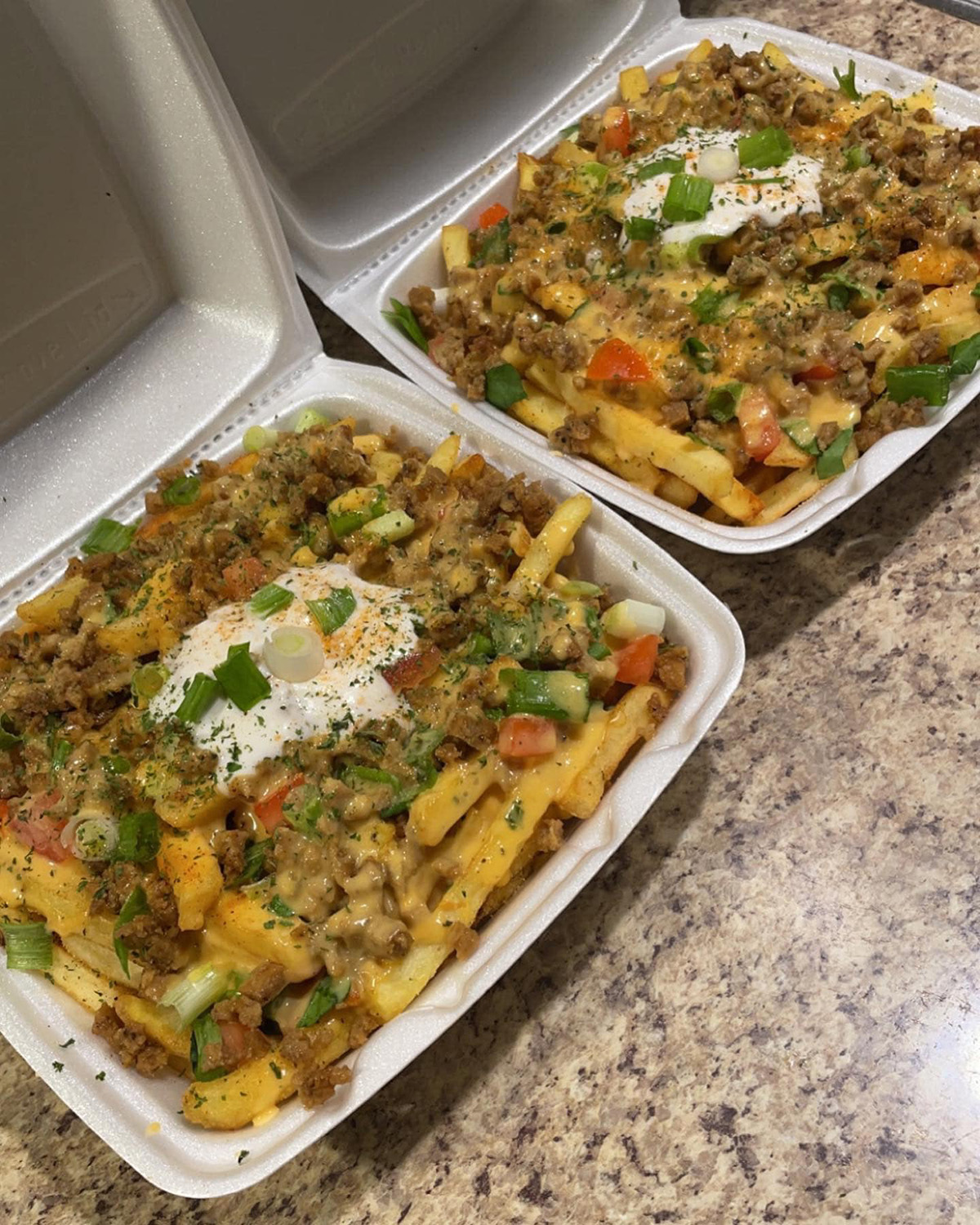 Two styrofoam take-out containers very full of french fries with toppings including vegan cheese, vegan meat, green onions, tomatoes, jalapenos, and spices. Photo by Lamiea Wilson.