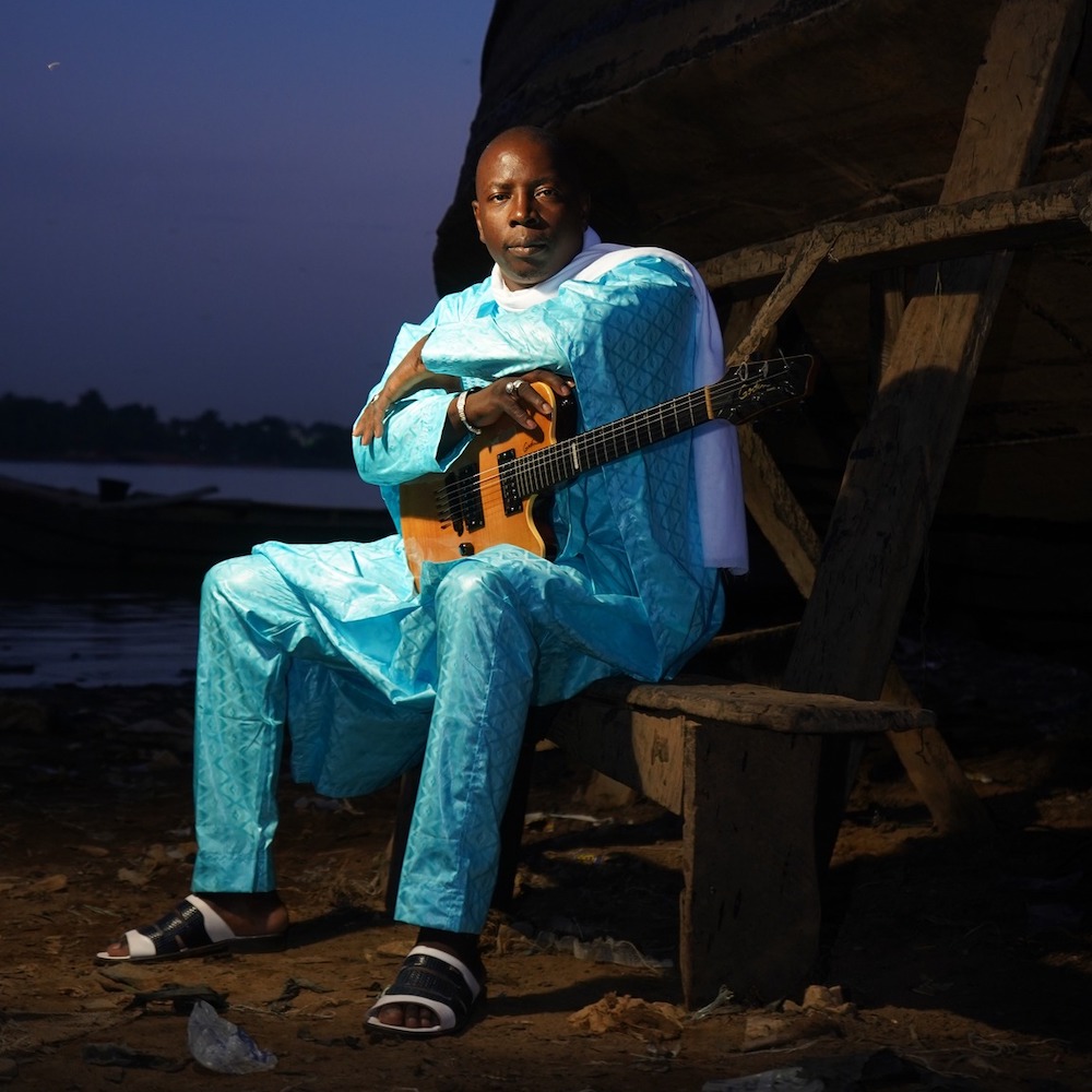 Vieux Farka Toure sits on a chair outside under the stars. He holds his guitar in his lap, wearing an emerald green outfit. 