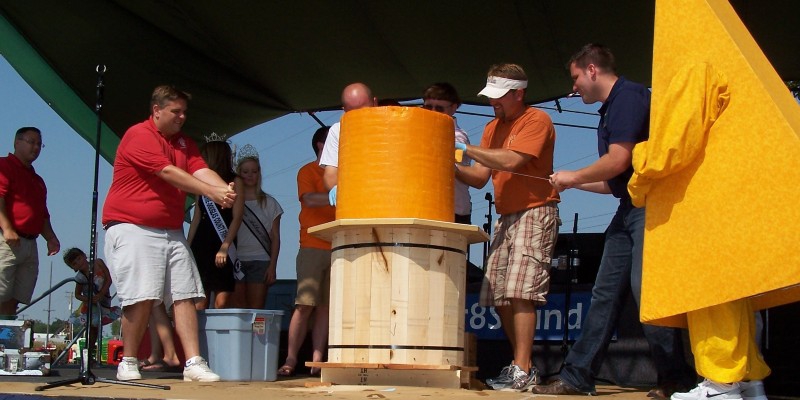 Men are gathered on a stage around a giant cylinder of cheese on a pedestal, using wires to cut it. Photo from Visit Champaign County website.