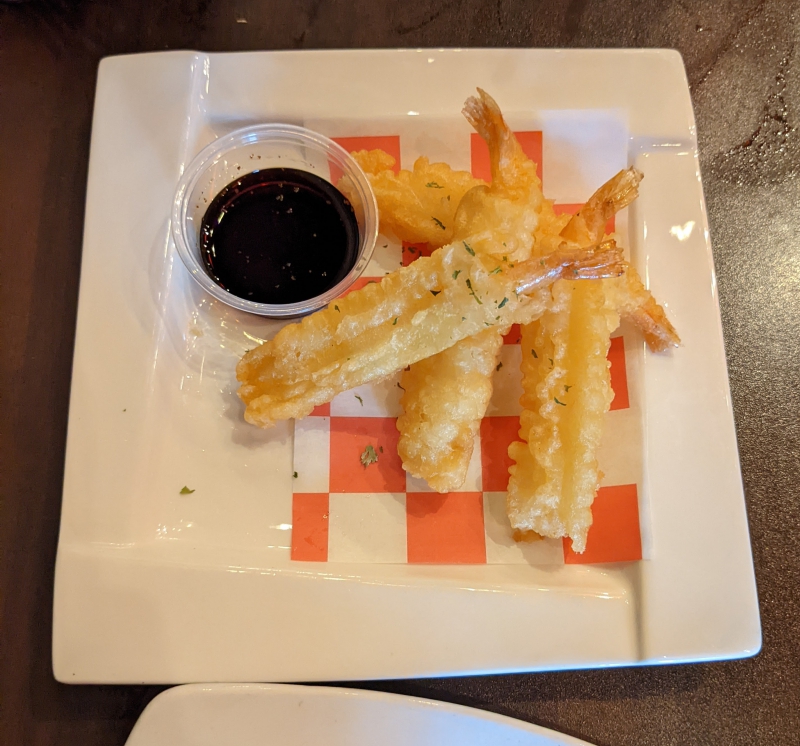  On a square white plate, five pieces of tempura shrimp are arranged on a red and white checkered paper. A small container of brown sauce sits next to them. Photo by Caitlin Aylmer.