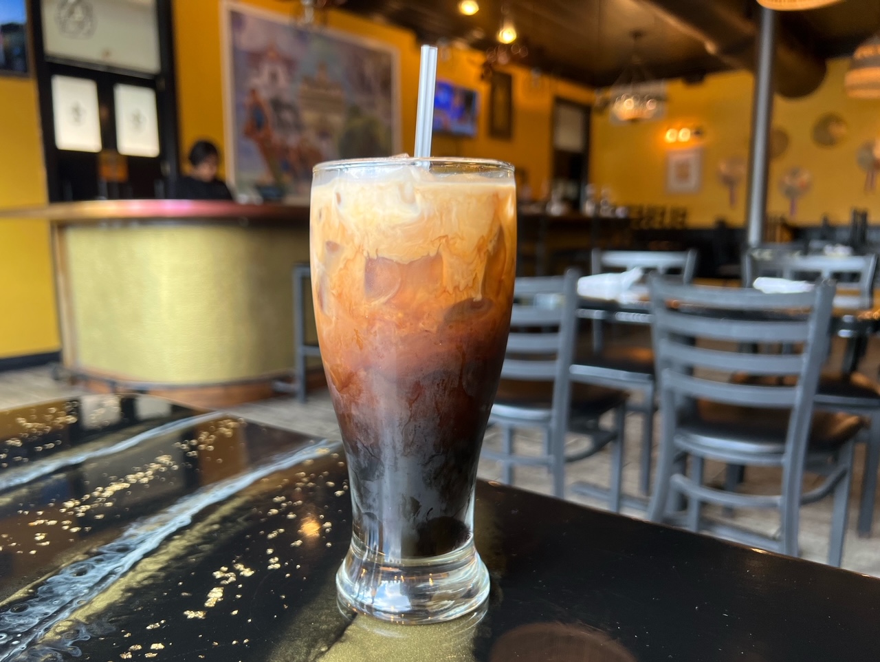 On a black table at Sticky Rice in Champaign, there is a tall glass of Thai iced coffee with swirled milk in the coffee. Photo by Alyssa Buckley.