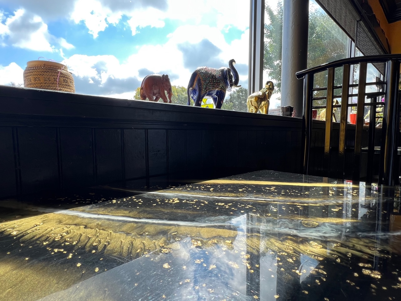 At Sticky Rice in Downtown Champaign, there are black and gold tables in a sun-drenched dining room. On the window ledge, there are various decorative elephants beside a window showing blue sky and wispy clouds. Photo by Alyssa Buckley.