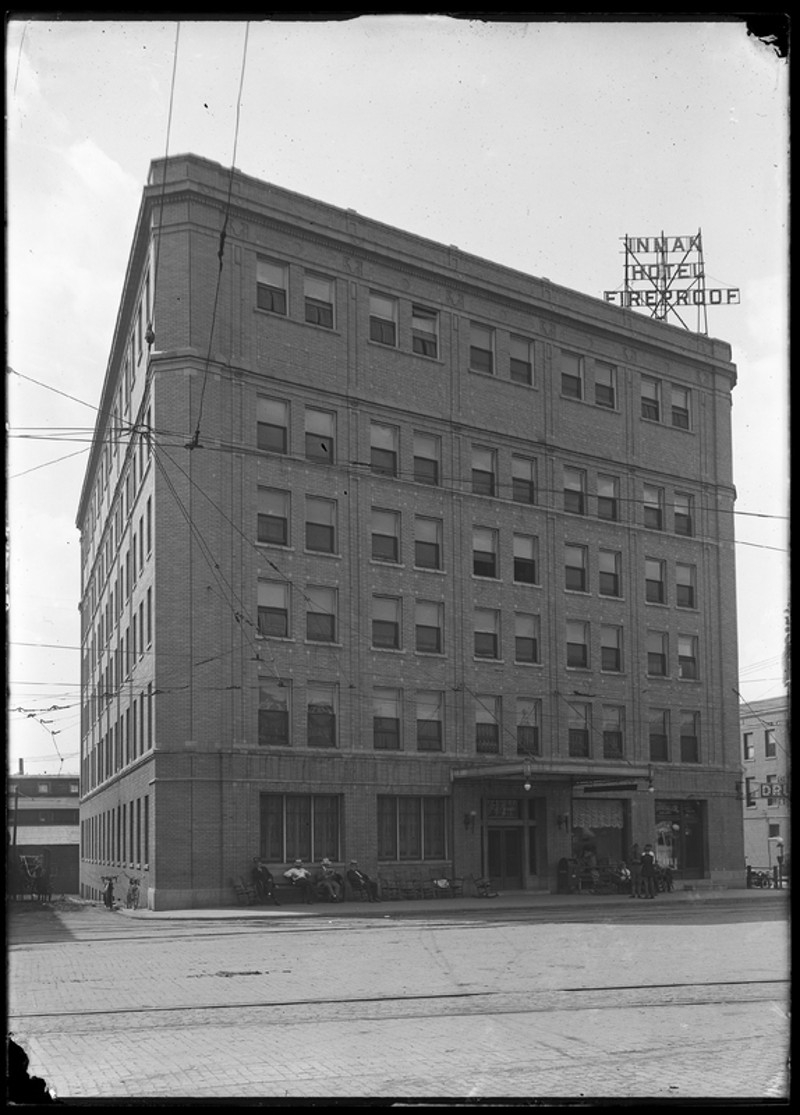 A black and white photo of a rectangular building with six floors, and a sign on the roof that says Inman Hotel Fireproof. It sits on a street corner, and there are several man sitting in front of the building. Photo from Champaign County Historical Archives.