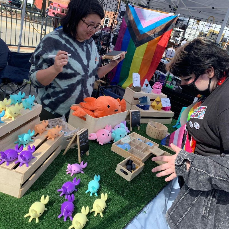A person is shopping at a vendor table covered in colorful crab figures. The vendor stands behind the table holding a small chalkboard and piece of chalk. Photo from Made Fest Facebook page.