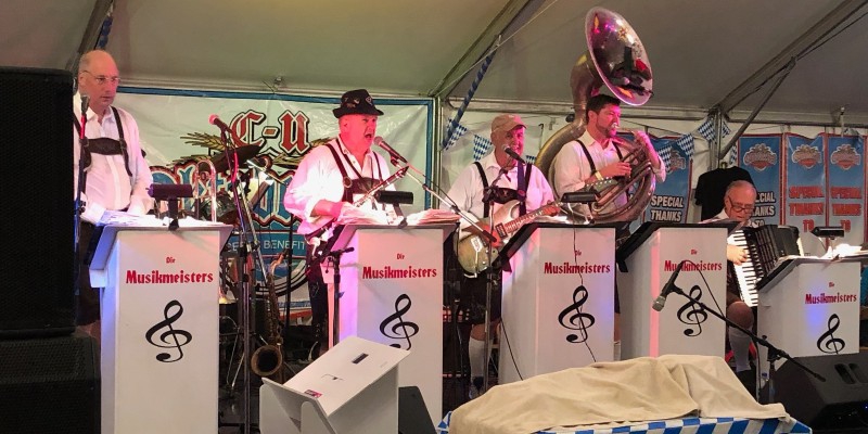 A row of musicians wearing lederhosen on a stage. They are behind band stands. Photo from C-U Oktoberfest Facebook page.