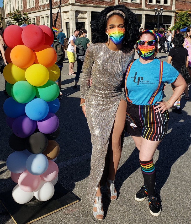 A drag queen in a long sparkling dress and long black hair is standing next to a person with a blue t-shirt and rainbow shorts with rainbow suspenders. They are both wearing rainbow masks. There is a balloon tower with the colors of the rainbow next to them. Photo from Uniting Pride Facebook page. 