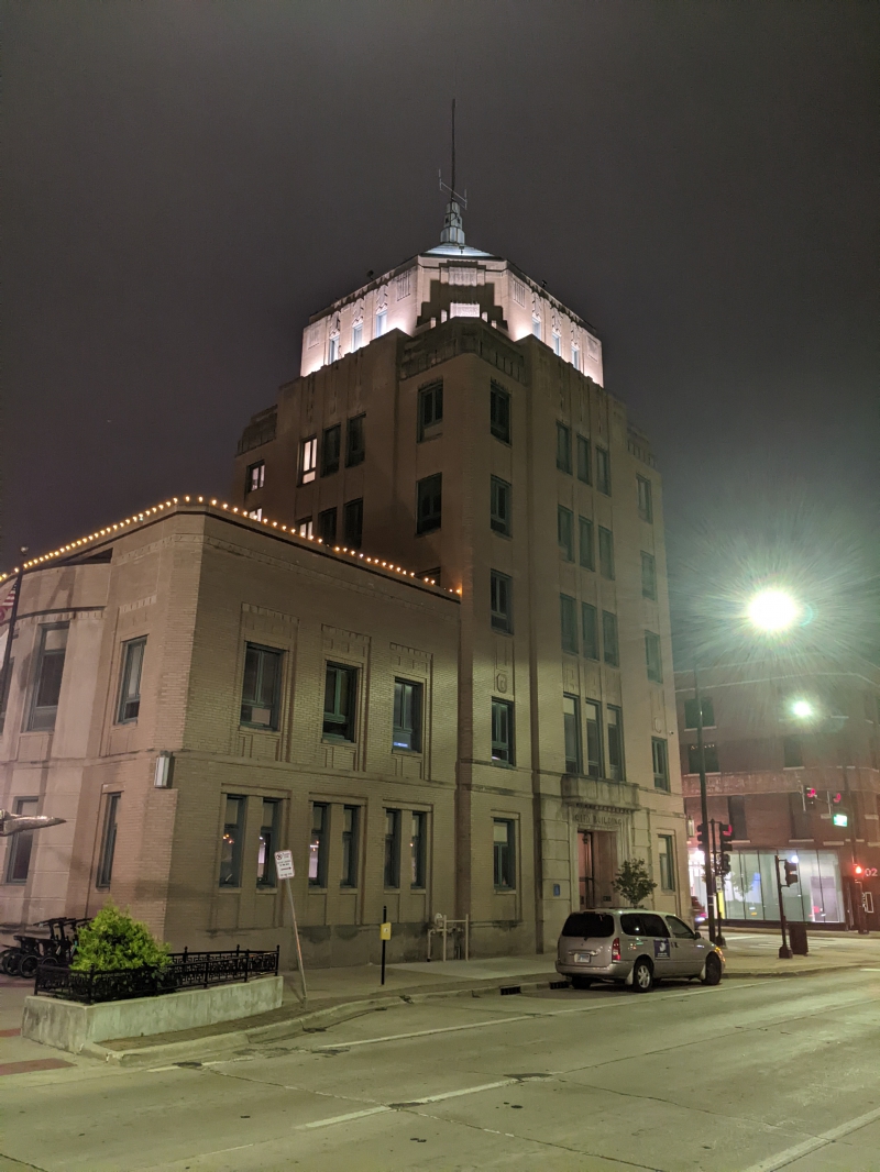 A beige brick building on a street corner. There is a lower, two story portion of the building and a taller 6 story portion with a greenish blue pointed roof. It is nighttime, and there are lights lining the lower roof, and flood lights illuminating the top of the tall portion. Photo by Tom Ackerman.