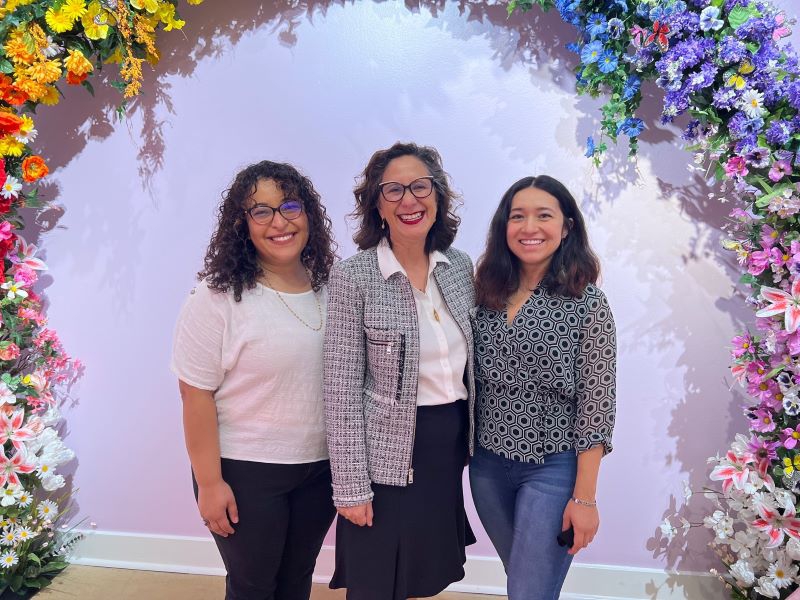 Three women are standing together and smiling under an archway of colorful flowers. Photo by Julie McClure.
