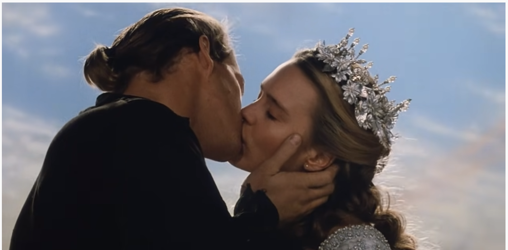 Cary Elwes, as Westley, pictured left, in all black, holds the face and is kissing Robin Wright, as Buttercup, pictured right. Wright wears a silver crown and a silver dress. Top image screenshot from the YouTube trailer.