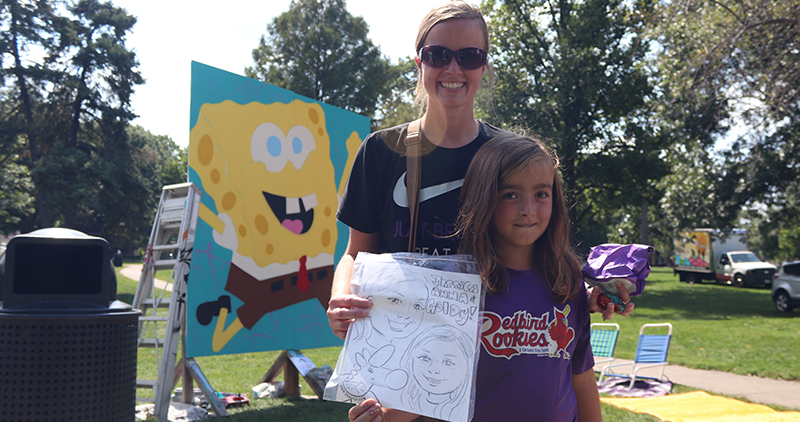 A mother and daughter standing at West Side Park holding a caricature drawing with a large painted canvas portrait of Sponge Bob Squarepants behind them.