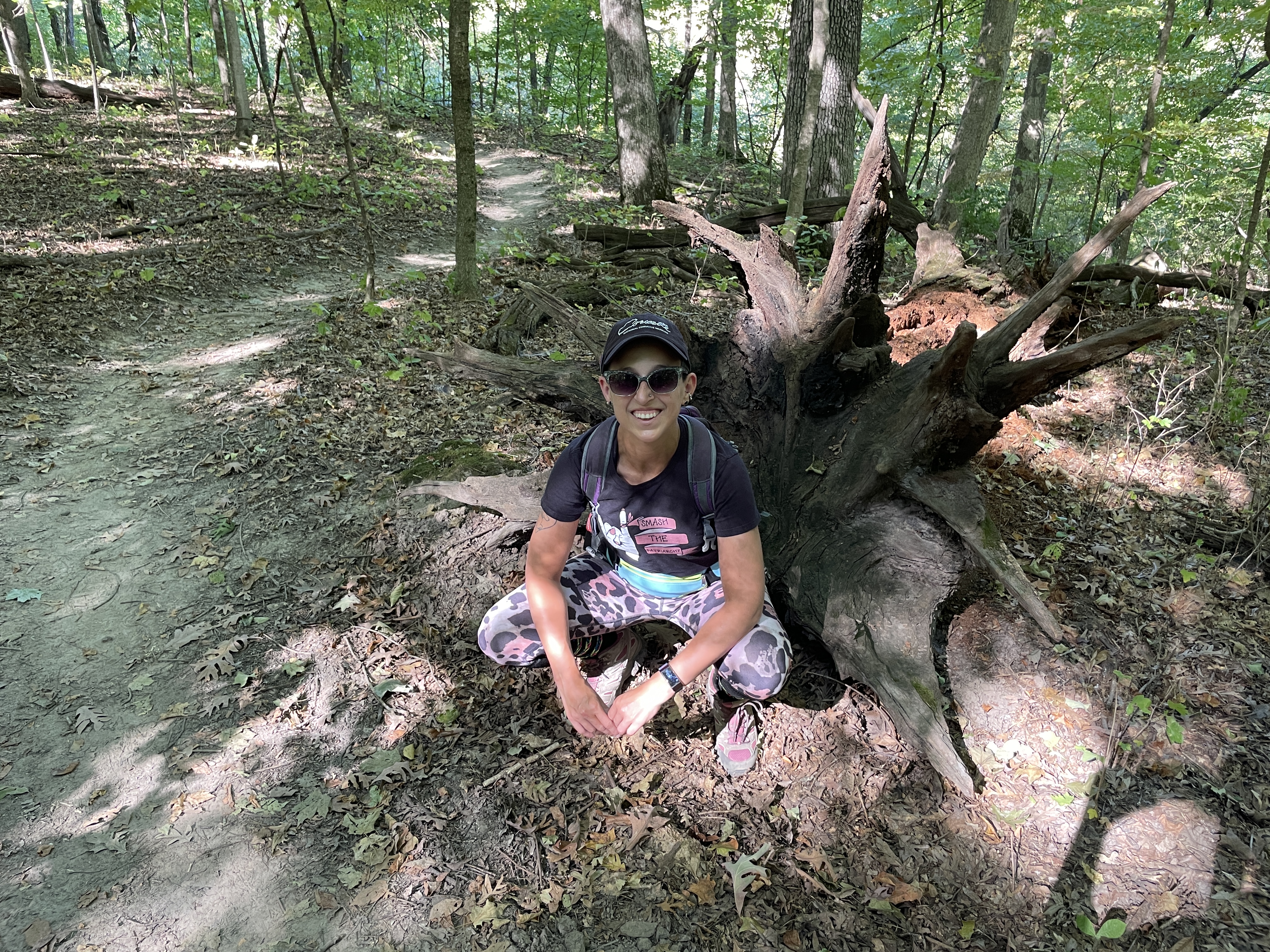 Photo the author, Mara, crouching in front of the roots of a fallen tree and next to the trail. Main colors in the photo are browns. 
