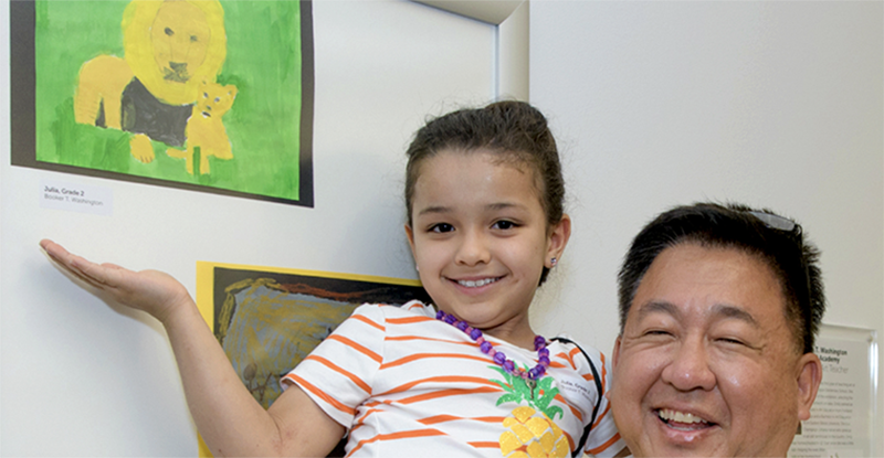 Photo of a young Asian girl held up by an older Asian male. Both are smiling as she points to her painting of a tiger hung on the wall.