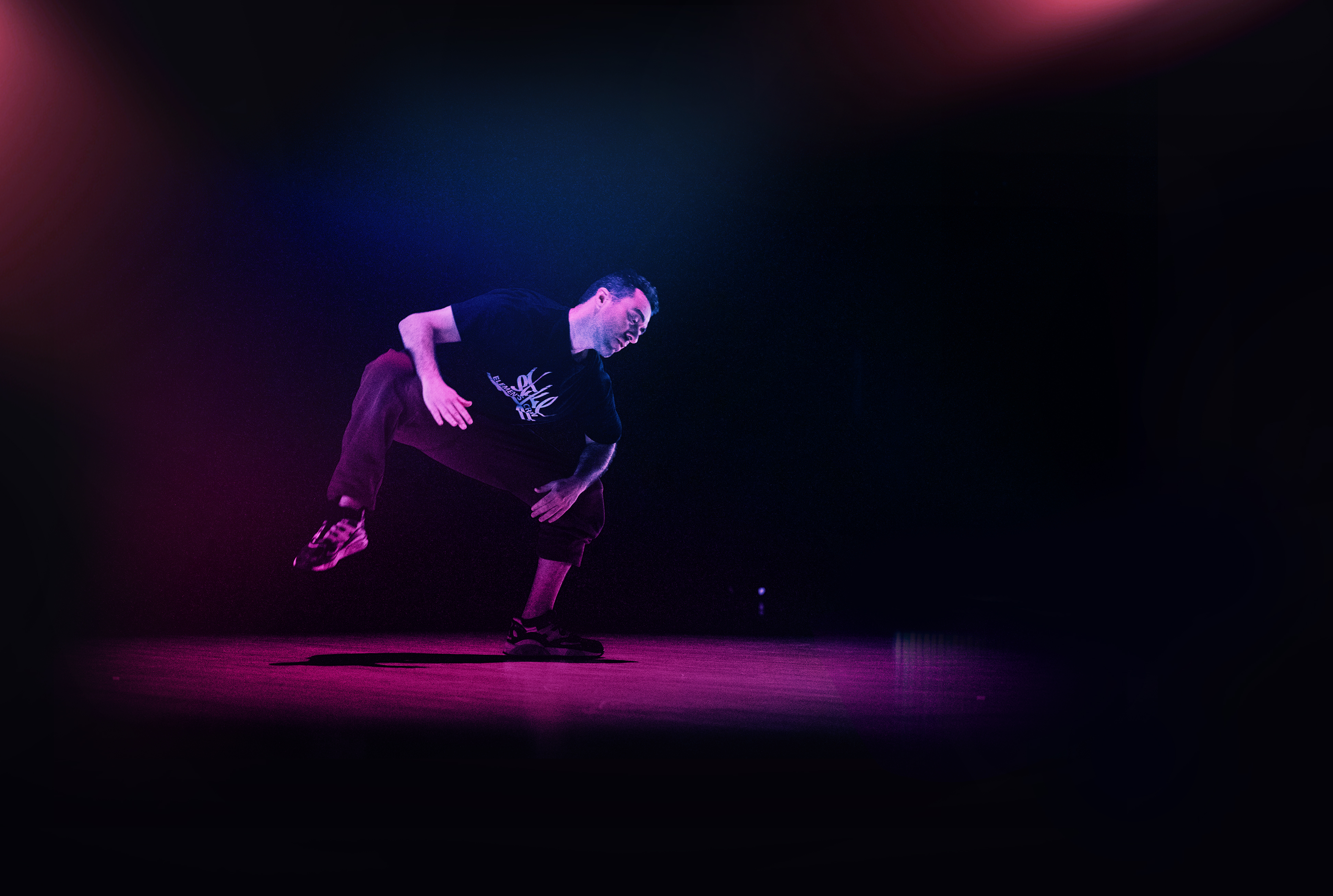  A male dancer is pictured mid-move, in a deep squat with his right leg lifted, so he is balanced on his left leg. He looks down and to the left. The stage area is dark, with a pink spotlight on him. Photo by Hoi Phan Do, courtesy of the Department of Dance.