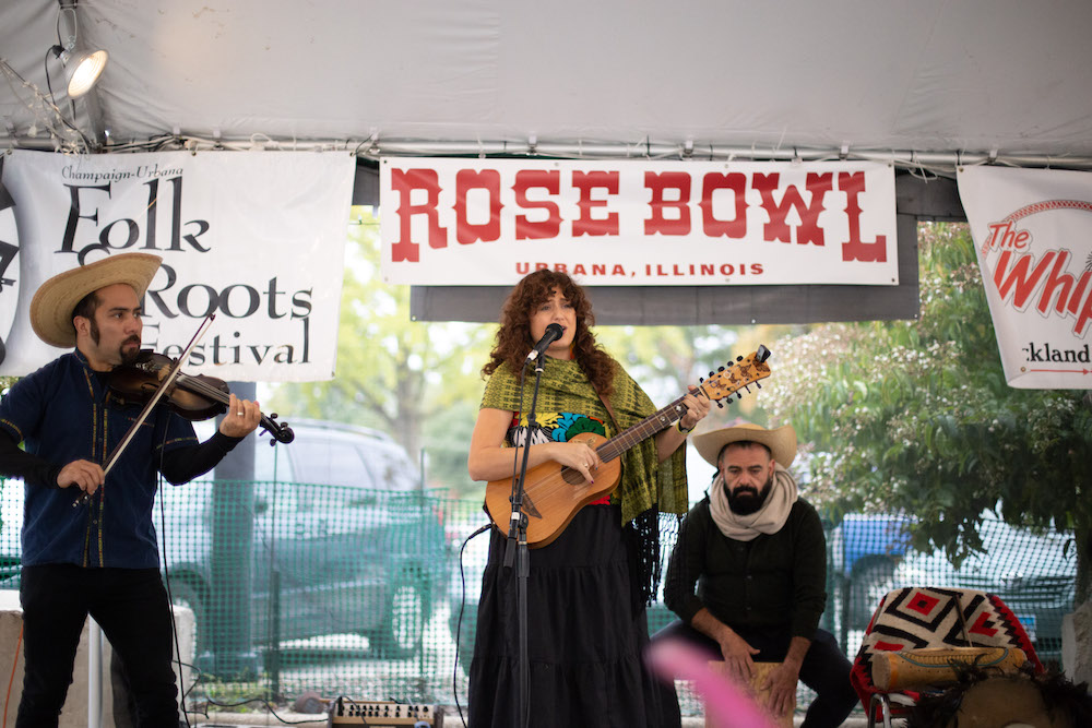 Three musicians play under the tent at Rose Bowl Tavern. On the left, a man plays fiddle. In the middle, a woman sings and plays a guitar. On the right, a man sits on a cajon drum and plays.