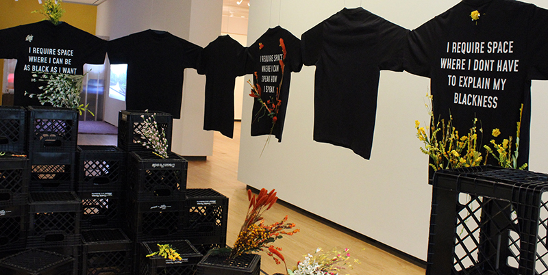 Stacks of black milk crates some filled with flowers, surrounded by black t-shirts linked arm in arm with sayings like 
