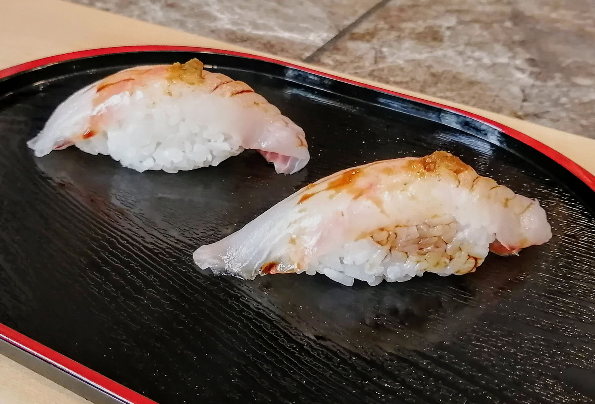  A close-up of two pieces of nigiri sushi with a dark brown sauce on top; the fish is white and the plate is black. Photo by Paul Young.