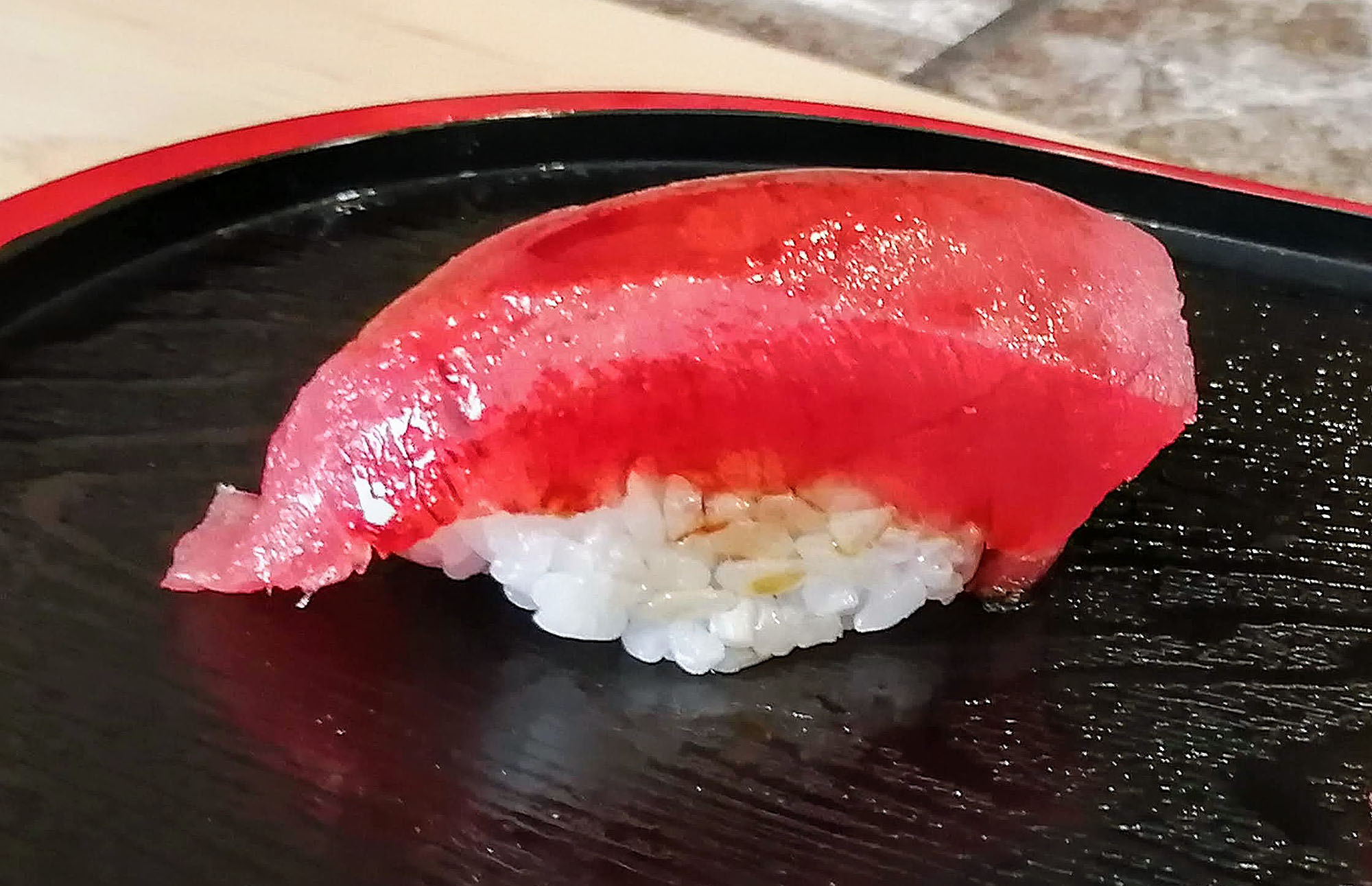 A close-up of a piece of nigiri sushi made with bright red raw tuna sitting on a black plate. Photo by Paul Young.