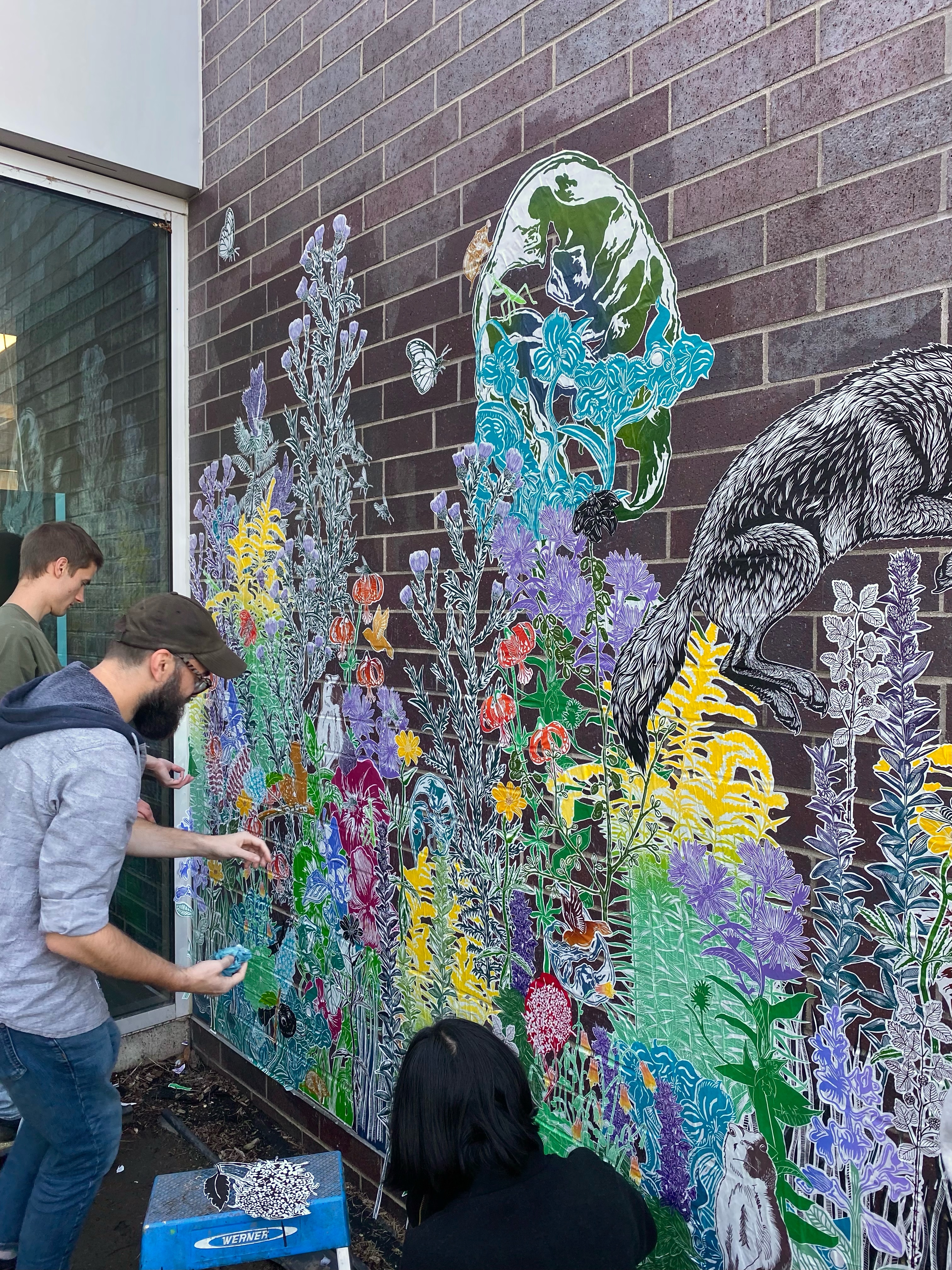 U of I students in ARTS 405 install an intricate paste-up mural on a brick wall. There are two young men to the left, holding pieces of paper to be pasted into the massive collage of plants. Photo by Emmy Lingscheit. 